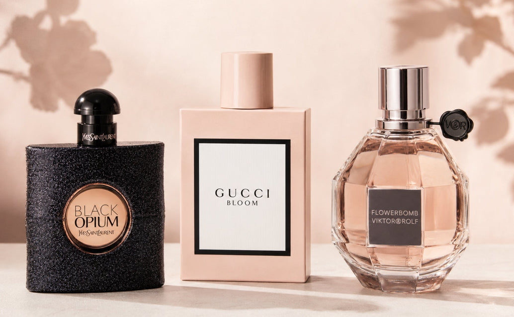 Three luxury perfume bottles—Black Opium, Gucci Bloom, and Flowerbomb—arranged on a matte ivory surface against a soft blush-beige background with delicate floral shadows, lit with diffused studio light in a minimalist campaign style.