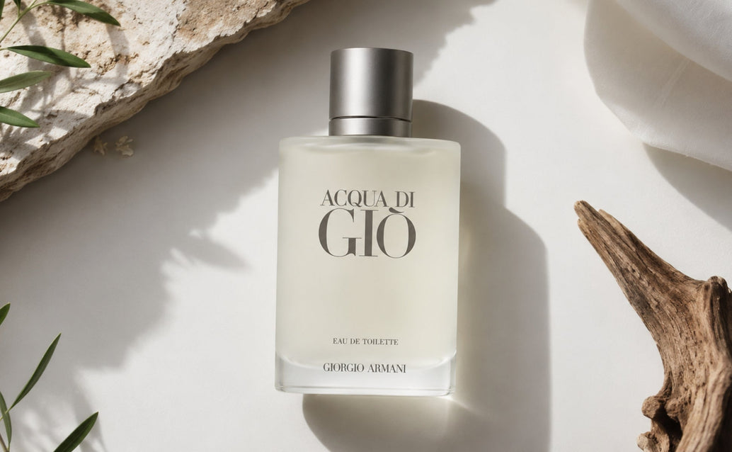 Acqua Di Giò by Giorgio Armani bottle photographed from above on a matte white surface with driftwood, green leaves, stone, soft fabric, and dramatic shadows.