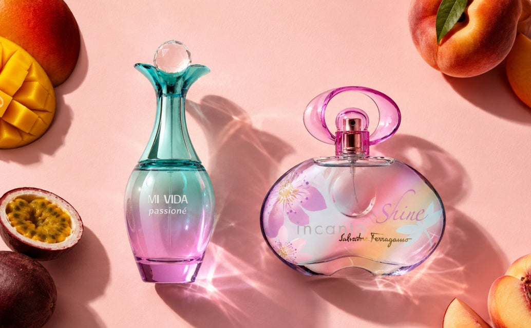 Two pastel perfume bottles, Mi Vida Passioné and Incanto Shine, arranged on a matte pink surface with mango, peach, and passionfruit in the corners, lit from the left to create soft shadows and sparkling glass reflections.