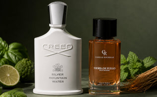 Creed Silver Mountain Water and Tierra de Fuego perfume bottles on a matte green surface with fresh green botanicals.