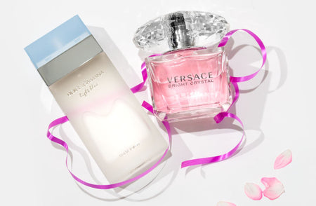 Two luxury perfume bottles on a white background: Dolce & Gabbana Light Blue on the left and Versace Bright Crystal on the right, accented with a pink ribbon and scattered rose petals.