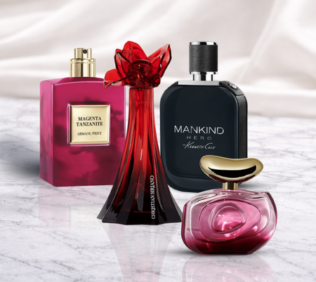 A group of four perfume bottles arranged on a marble surface, including a pink square bottle labeled “Magenta Tanzanite,” a tall red sculptural bottle, a dark blue “Mankind Hero” bottle, and a small rounded pink bottle with a gold cap, set against a soft white fabric background.
