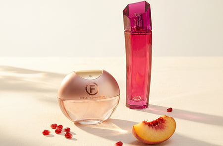 Two pink perfume bottles, including a round French Connection bottle, displayed on a sunlit cream surface beside a sliced peach and scattered pomegranate seeds.