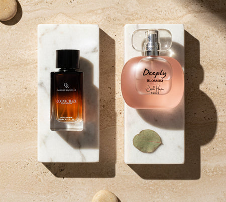 Two perfume bottles displayed on white marble blocks against a beige stone background, with sunlight, shadows, and a small green leaf