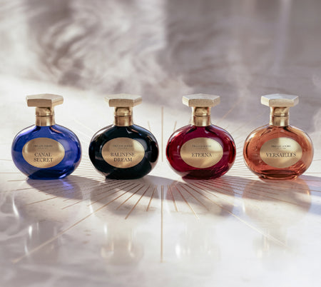 Four ornate perfume bottles in blue, black, red, and rose-gold displayed side by side on a luminous marble surface with a soft, elegant background.